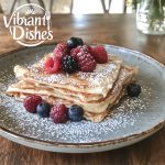 Stack of golden crepes garnished with berries and powdered sugar.